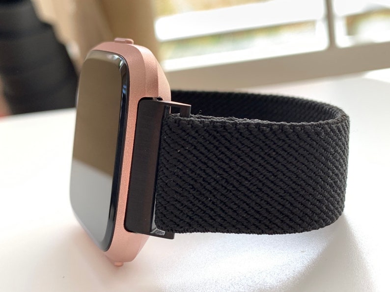May include: A rose gold smartwatch with a black elastic band. The watch face is black and rectangular.
