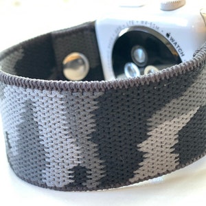 May include: A black and grey camouflage patterned Apple Watch band with a silver buckle. The band is made of a soft, stretchy material.