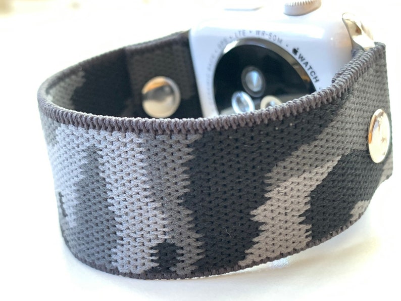 May include: A black and grey camouflage patterned Apple Watch band with a silver buckle. The band is made of a soft, stretchy material.