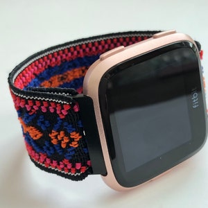 May include: A rose gold Fitbit smartwatch with a black and white patterned elastic band. The band has red, orange, and blue accents.