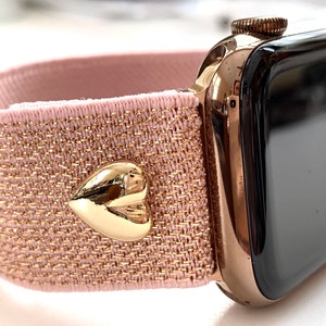 May include: A rose gold Apple Watch with a pink and gold glitter elastic band. The band has a gold heart-shaped charm.