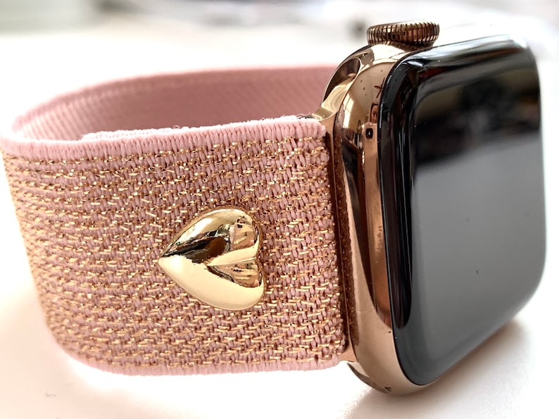 May include: A rose gold Apple Watch with a pink and gold glitter elastic band. The band has a gold heart-shaped charm.