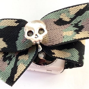 May include: A camouflage patterned Apple Watch band with a silver skull charm. The watch face is visible and says "Ceramic Case Sapphire 32mm".