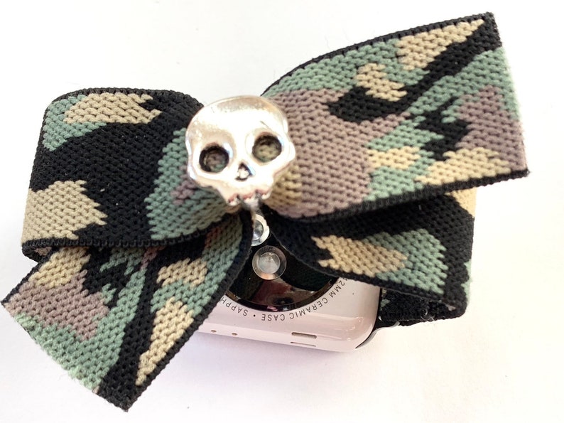 May include: A camouflage patterned Apple Watch band with a silver skull charm. The watch face is visible and says "Ceramic Case Sapphire 32mm".