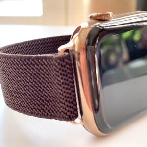 Puede incluir: Un Apple Watch dorado con una banda deportiva de bucle marr&oacute;n. La esfera del reloj es negra y la banda est&aacute; hecha de un material suave y tejido.