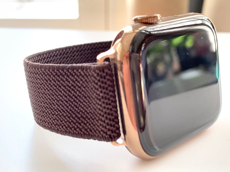 Puede incluir: Un Apple Watch dorado con una banda deportiva de bucle marr&oacute;n. La esfera del reloj es negra y la banda est&aacute; hecha de un material suave y tejido.