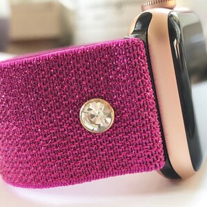 May include: A pink glitter watch band with a clear rhinestone accent. The band is compatible with Apple Watch.