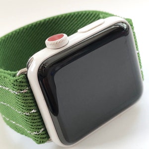 Puede incluir: Un Apple Watch blanco con una esfera negra y una banda deportiva verde. La banda tiene una franja blanca que recorre el centro.