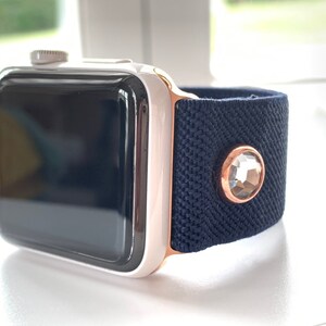 Puede incluir: Un Apple Watch blanco con una banda de oro rosa y una banda elástica azul marino con un acento de cristal transparente.