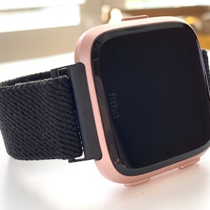 May include: A rose gold Fitbit smartwatch with a black woven band. The watch face is black and displays the word "fitbit".