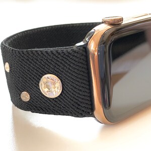 May include: A rose gold Apple Watch with a black sport band featuring small rhinestones. The watch face is black and the band is made of a textured fabric.