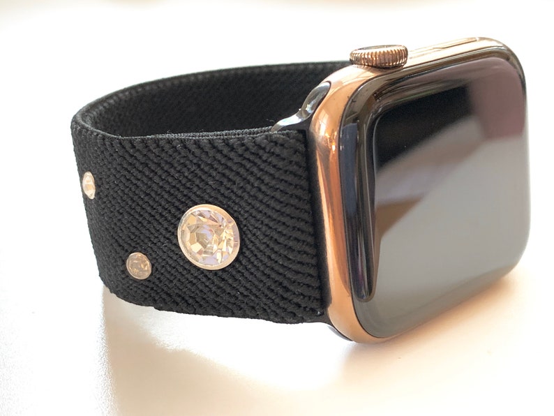 May include: A rose gold Apple Watch with a black sport band featuring small rhinestones. The watch face is black and the band is made of a textured fabric.
