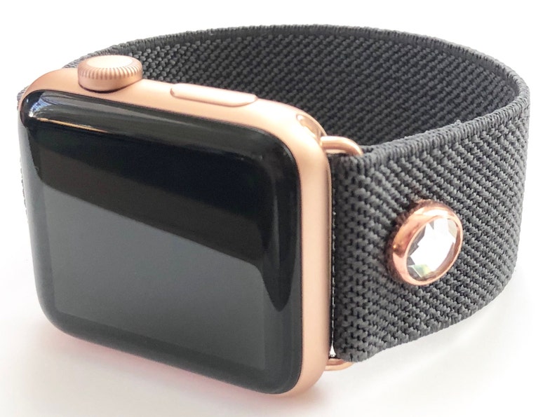 May include: A rose gold Apple Watch with a black face and a gray elastic band with a single rhinestone accent.