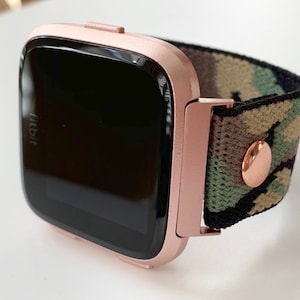 May include: Rose gold Fitbit smartwatch with a camouflage patterned band. The watch face is black and the band is made of a soft, stretchy material.