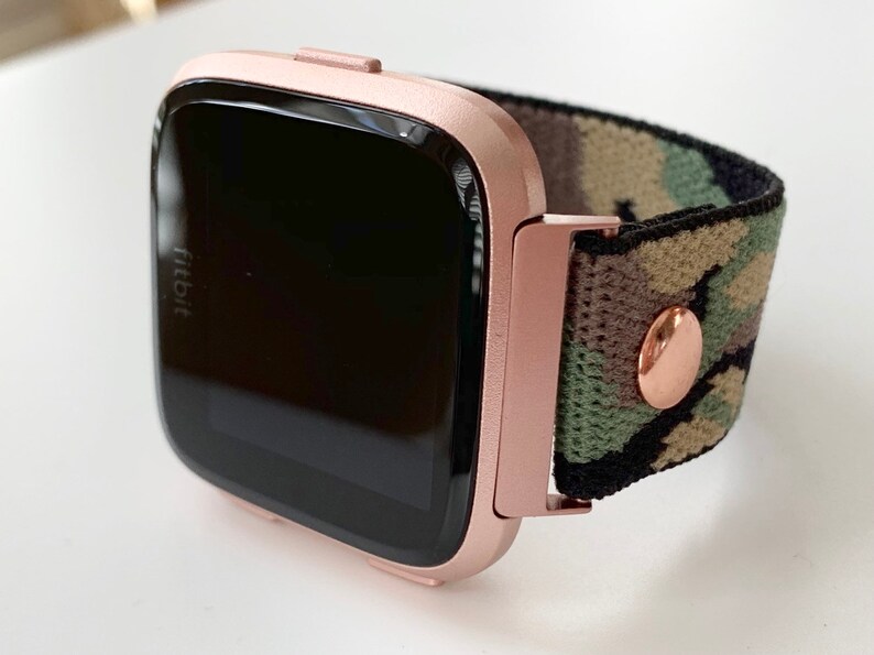 May include: Rose gold Fitbit smartwatch with a camouflage patterned band. The watch face is black and the band is made of a soft, stretchy material.