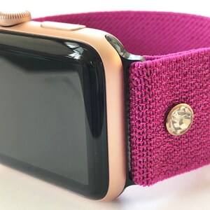 May include: A rose gold Apple Watch with a pink sparkly fabric band. The band has a small clear rhinestone on it.