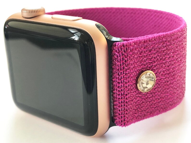 May include: A rose gold Apple Watch with a pink sparkly fabric band. The band has a small clear rhinestone on it.