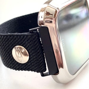 May include: A silver metal case with a black elastic band and a silver button. The case has a clear screen with the word "Fitbit" visible.