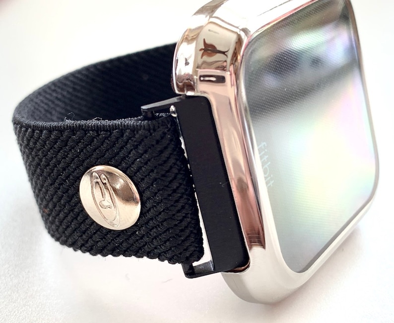 May include: A silver metal case with a black elastic band and a silver button. The case has a clear screen with the word "Fitbit" visible.