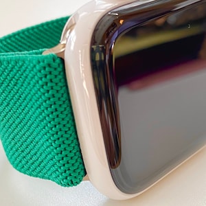 K&ouml;nnte beinhalten: Eine wei&szlig;e Apple Watch mit einem gr&uuml;nen Sportarmband. Das Zifferblatt der Uhr ist schwarz und das Armband besteht aus einem weichen, dehnbaren Material.