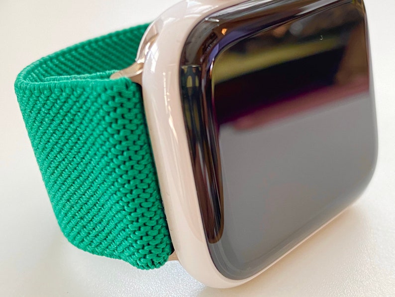 K&ouml;nnte beinhalten: Eine wei&szlig;e Apple Watch mit einem gr&uuml;nen Sportarmband. Das Zifferblatt der Uhr ist schwarz und das Armband besteht aus einem weichen, dehnbaren Material.
