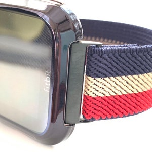 Puede incluir: Un reloj inteligente Fitbit negro con una banda el&aacute;stica a rayas azul marino, blanco y rojo. La banda tiene una textura tejida.