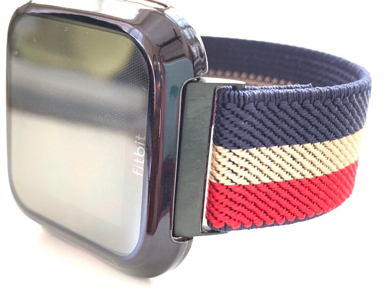 Puede incluir: Un reloj inteligente Fitbit negro con una banda el&aacute;stica a rayas azul marino, blanco y rojo. La banda tiene una textura tejida.