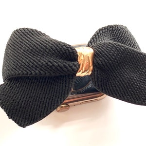 May include: A black fabric bow with a gold metal accent is attached to a gold Apple Watch.