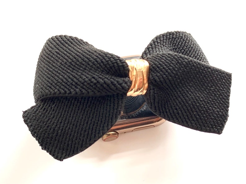 May include: A black fabric bow with a gold metal accent is attached to a gold Apple Watch.