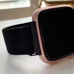 May include: A rose gold Fitbit smartwatch with a black woven band. The watch face is black and the Fitbit logo is visible on the watch face.
