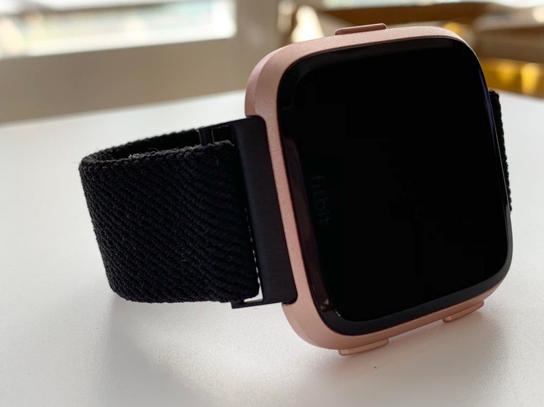 May include: A rose gold Fitbit smartwatch with a black woven band. The watch face is black and the Fitbit logo is visible on the watch face.