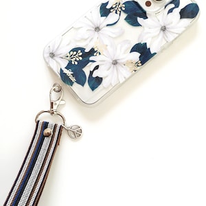 May include: A clear phone case with a floral design featuring white flowers and dark blue leaves. The case has a gold accent and the word "Sonix" printed on it. A striped phone strap with a peace sign charm is attached to the case.