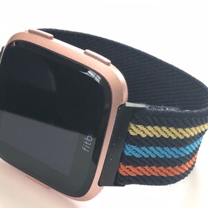 May include: A rose gold Fitbit smartwatch with a black and multi-colored striped elastic band. The watch face is black and displays the word "Fitbit".