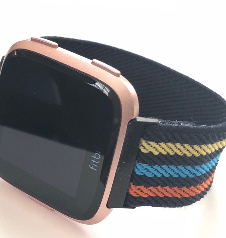 May include: A rose gold Fitbit smartwatch with a black and multi-colored striped elastic band. The watch face is black and displays the word "Fitbit".
