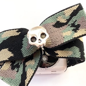 May include: A camouflage patterned fabric bow with a silver skull charm attached to it. The bow is wrapped around a white Apple Watch with the text "Ceramic Case 42mm" on the side.