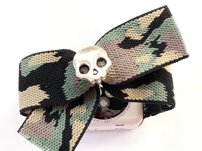 May include: A camouflage patterned fabric bow with a silver skull charm attached to it. The bow is wrapped around a white Apple Watch with the text "Ceramic Case 42mm" on the side.