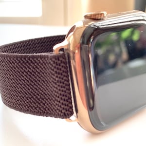 Puede incluir: Un Apple Watch de oro rosa con una banda deportiva de bucle marr&oacute;n. La esfera del reloj es negra y la banda est&aacute; hecha de un material suave y el&aacute;stico.