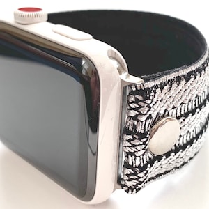 Puede incluir: Un Apple Watch blanco con una banda de patr&oacute;n negro y plateado. La banda tiene un bot&oacute;n de metal plateado.