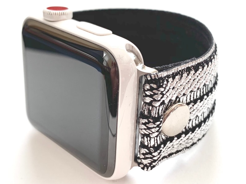 Puede incluir: Un Apple Watch blanco con una banda de patr&oacute;n negro y plateado. La banda tiene un bot&oacute;n de metal plateado.