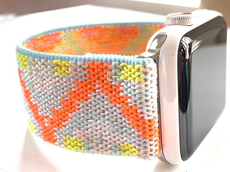 May include: A colorful knitted watch band in shades of orange, yellow, blue, and white. The band is attached to a white Apple Watch.