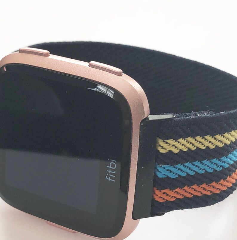 May include: A rose gold Fitbit smartwatch with a black and multi-colored striped band. The watch face is black and displays the word "Fitbit".