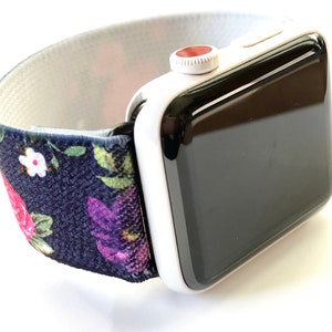 May include: A white Apple Watch with a black face and a floral patterned elastic band. The band is navy blue with pink, purple, and green flowers.