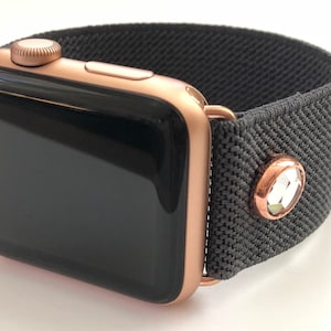 May include: A rose gold Apple Watch with a black screen and a gray fabric band with a rose gold crystal accent.
