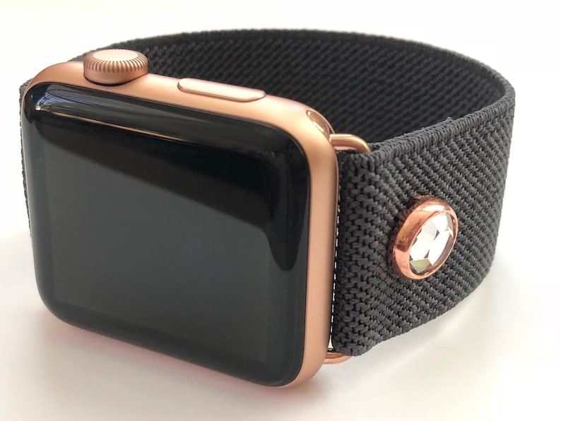 May include: A rose gold Apple Watch with a black screen and a gray fabric band with a rose gold crystal accent.