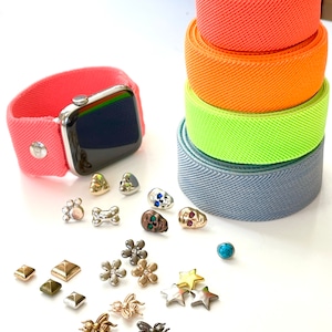 Könnte beinhalten: Ein Stapel bunter elastischer Uhrenbänder in Orange-, Gelb-, Grün- und Grautönen. Eine silberne Apple Watch mit einem rosafarbenen Band ist neben dem Stapel abgebildet.  Eine Vielzahl kleiner Charms sind um die Uhr und die Bänder verstreut, darunter Sterne, Blumen, Totenköpfe und Bienen.