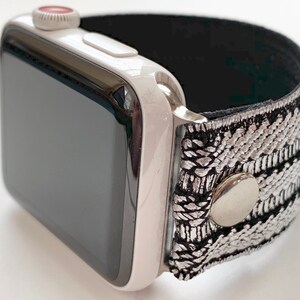 Puede incluir: Un Apple Watch blanco con una banda de patr&oacute;n negro y plateado. La banda tiene un bot&oacute;n plateado.