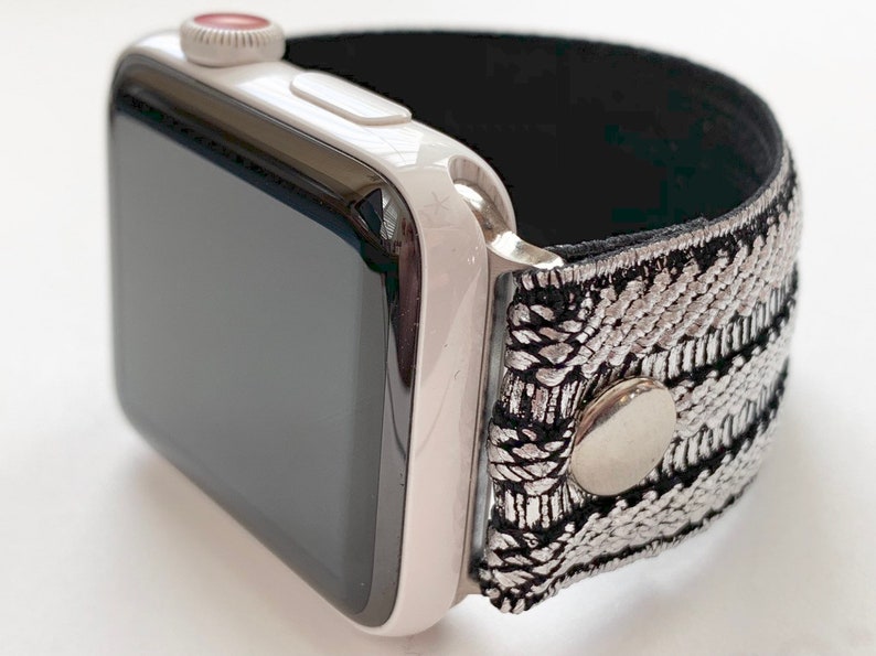 Puede incluir: Un Apple Watch blanco con una banda de patr&oacute;n negro y plateado. La banda tiene un bot&oacute;n plateado.