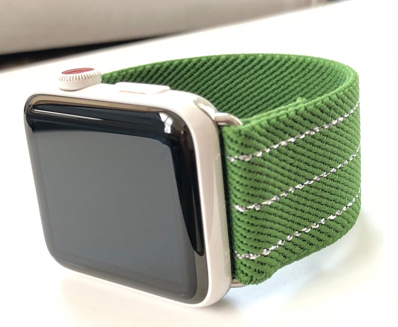 Puede incluir: Un Apple Watch plateado con una banda de tela verde. La banda tiene un detalle de costura blanca.