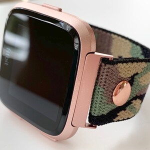 May include: Rose gold Fitbit smartwatch with a camouflage patterned band. The band has a rose gold button.