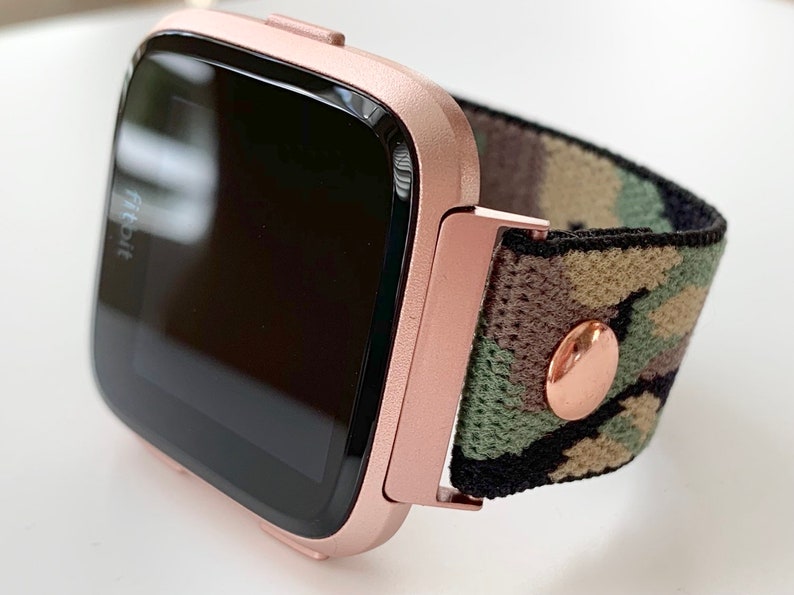 May include: Rose gold Fitbit smartwatch with a camouflage patterned band. The band has a rose gold button.